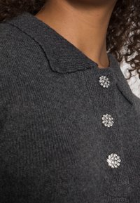 Pull gris en maille avec un tissu texturé, un petit col et trois boutons décoratifs en rhinestone au niveau de l'encolure.