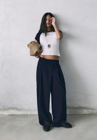 Mladá žena v bielom a čiernom krátkom topu a širokých čiernych nohaviciach stojí opretá o sivú stenu, pričom jednou rukou drží zloženú béžovú bundu.