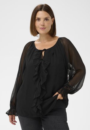 Femme portant un chemisier noir avec des manches longues transparentes et un détail froncé à l'avant, posant avec une main dans sa poche sur un fond clair.