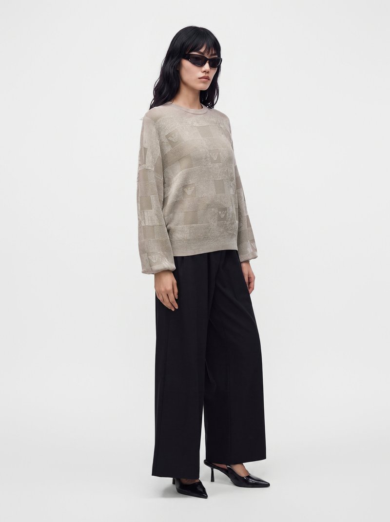 Femme portant un pull beige à motifs surdimensionné, un pantalon noir large, des talons noirs et des lunettes de soleil foncées, debout sur un fond blanc uni.