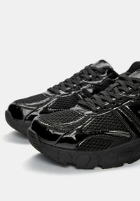 Pantofi sport negri cu o textură din plasă și accente din piele lacuită strălucitoare. Au tălpi din cauciuc și șireturi negre, concepuți pentru confort și suport.
