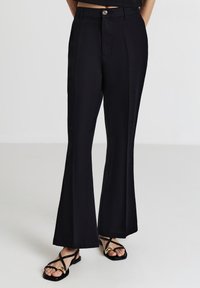 Schwarze, ausgestellte Hosen mit glatter Textur, ausgestattet mit einem Knopfverschluss vorne, Seitentaschen und dezenten Faltendetails.