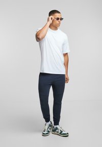 Hvit bomull T-skjorte, marineblå joggebukser med kontrastpaneler og grønne/hvite joggesko. Firkantede solbriller fullfører det uformelle antrekket.