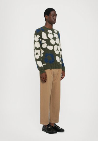 Jersey verde de punto con patrones abstractos en blanco y azul, combinado con pantalones beige. El modelo lleva zapatos negros y está de pie frente a un fondo blanco.