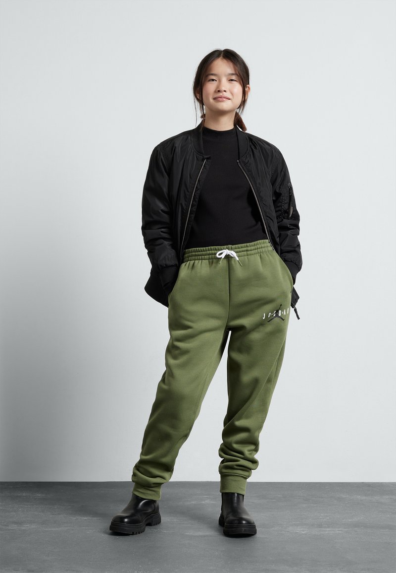Giacca bomber nera sopra un top a costine nero, abbinata a pantaloni alla zuava verde oliva con coulisse bianca e dettagli testuali. Stivali neri spessi.