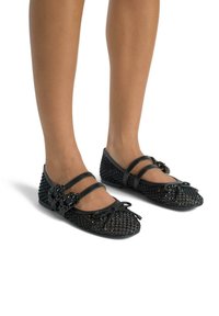 Bailarinas negras hechas de material texturizado con un diseño de dos correas, lazos decorativos y adornos brillantes en la punta.