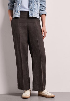 Osoba ubrana w ciemne, szerokie spodnie, białe trampki, biały top i niebieską kurtkę dżinsową z podwiniętymi rękawami, stojąca przy płaskiej ścianie.