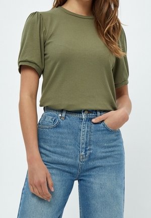 Kobieta ubrana w oliwkowozielony prążkowany top z krótkimi bufkowatymi rękawami i wysokim stanem dżinsy w niebieskim kolorze, z jedną ręką w kieszeni.
