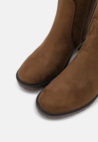Botas de tobillo de ante marrón con punta redonda y cremallera lateral, que cuentan con una superficie texturizada y suela de goma para mayor agarre.