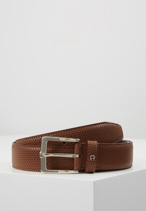 Ceinture en cuir brun texturé avec boucle rectangulaire argentée, roulée sur une surface blanche sur un fond clair uni.