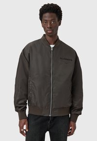 Brun bomberjakke med lynlås, ribbede manchetter og kant, samt sort logo tekst. Glat tekstur, let design og afslappet pasform.