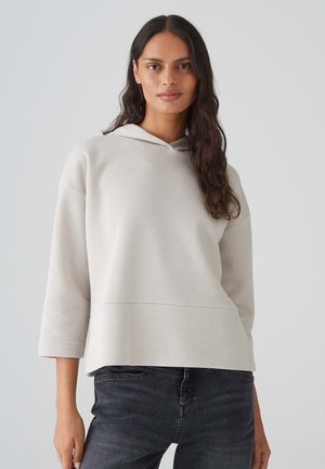 Felpa grigia chiara con cappuccio, caratterizzata da una vestibilità rilassata, maniche a tre quarti e una texture liscia. Indossata con jeans scuri.