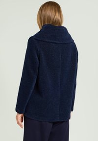 Kobieta o długaśnych, jasnobrązowych włosach, ubrana w ciemnoniebieską, teksturowaną kurtkę z długim rękawem, stojąca tyłem do aparatu.