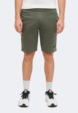 Homme portant un short Puma vert olive, des chaussettes blanches et des baskets blanches Puma, debout les mains détendues le long du corps.