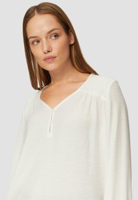Blouse blanche à manches longues avec décolleté en V, présentant un plastron avec des boutons et un tissu texturé. Fronces subtiles aux épaules.