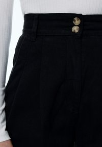 Pantalones negros de cintura alta con un tejido texturizado, que presentan un frente plano, bolsillos laterales y dos grandes botones claros en la cintura.
