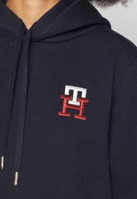 Felpa con cappuccio blu navy con un logo ricamato bianco e rosso sul lato sinistro del petto. Ha un cappuccio con coulisse e tessuto testurizzato.