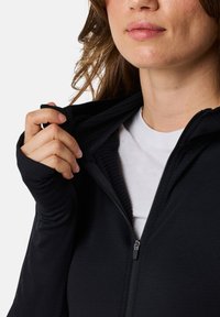 Svart zip-up-jacka med en strukturerad innervaddering, figurnära ärmar och en slät utsida. Dragkedjan är delvis öppen.