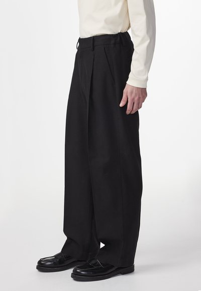 Man in zwarte wijde broek, zwarte instappers en een off-white shirt met lange mouwen, staande tegen een effen witte achtergrond.