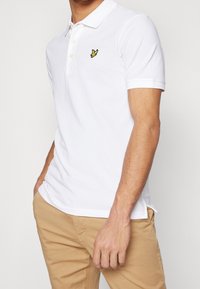 Polo en coton blanc avec col côtelé, patte à trois boutons et petit logo brodé jaune sur la poitrine gauche.