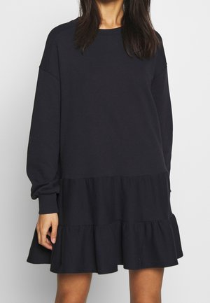 Frau trägt ein schwarzes Kleid mit langen Ärmeln und gestuftem Rüschensaum, steht vor einem einfachen hellen Hintergrund.