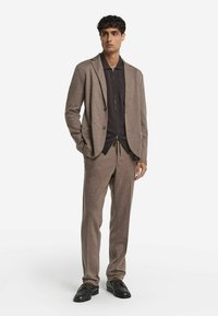 Dopasowany, jasnobrązowy garnitur o teksturowanym wykończeniu, z ciemną koszulą zapinaną na zamek błyskawiczny pod spodem oraz czarnymi sznurowanymi butami. Dopasowany krój.