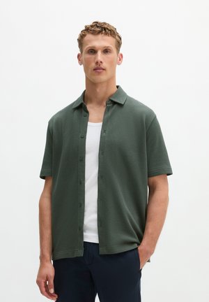 Mann trägt ein dunkelgrünes, kurzärmeliges, strukturiertes Hemd mit Knöpfen über einem weißen Tanktop und dunkler Hose, steht vor einem schlichten weißen Hintergrund.
