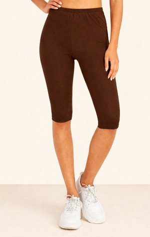 Femme portant des leggings marron longueur genou et des chaussures de sport blanches, debout avec une main sur la hanche et une main relâchée.