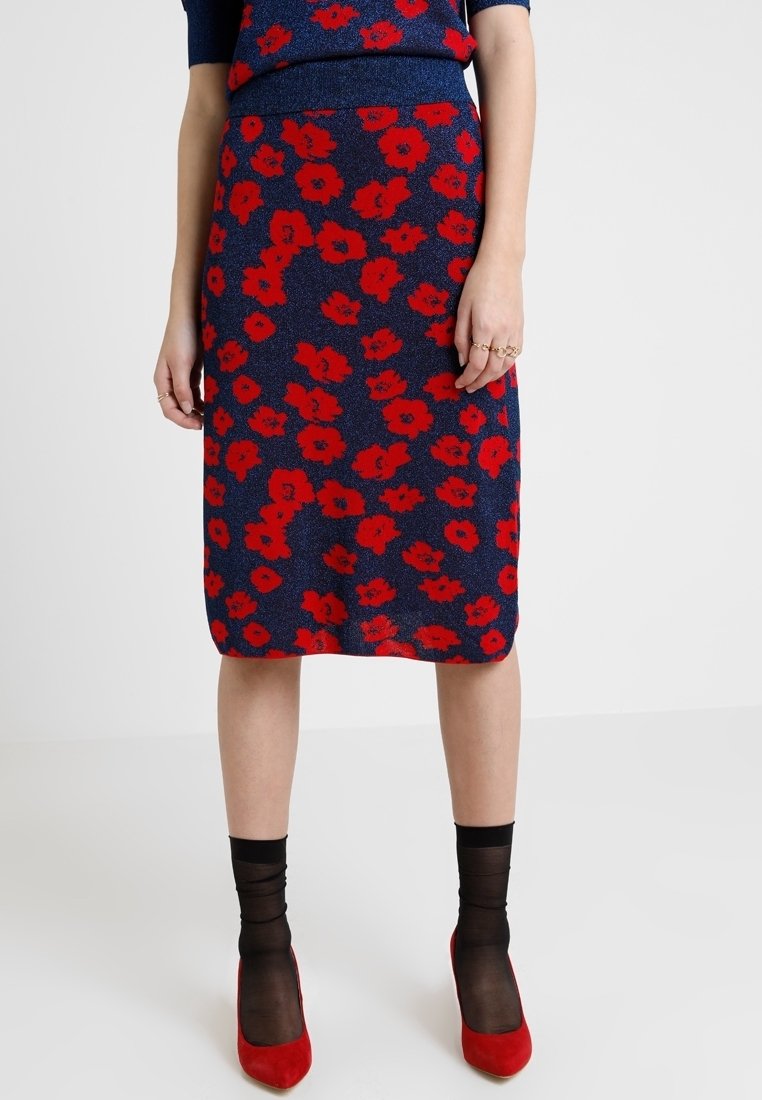 Jupe bleu marine à motif floral rouge portée avec des socquettes noires translucides et des talons pointus rouges, mains relâchées le long du corps.