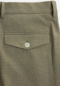 Textured olive green fabric of pants featuring a back pocket with a button closure. The design includes a subtle waffle pattern.