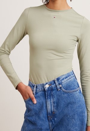 Woman wearing light green long-sleeve fitted shirt tucked into high-waisted blue denim jeans with one hand in pocket.