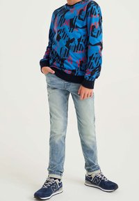 Ragazzo che indossa una maglietta a maniche lunghe blu, rossa e nera a trama, jeans azzurri chiari e sneaker blu navy, in piedi con una mano in tasca.