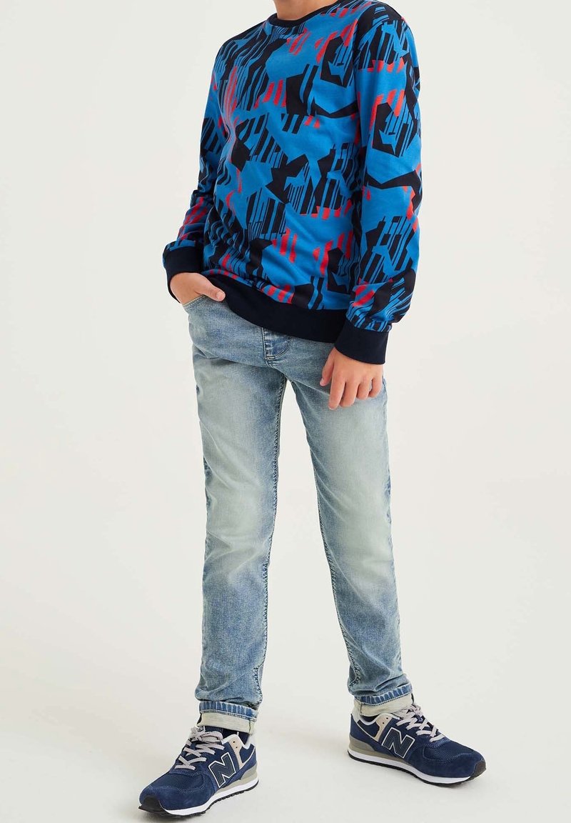 Ragazzo che indossa una maglietta a maniche lunghe blu, rossa e nera a trama, jeans azzurri chiari e sneaker blu navy, in piedi con una mano in tasca.