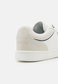 Bijeli tenisice s glatkom kožom i suede završnom obradom, s diskretnim reljefnim Levi's logotipom, bijelim gumenim potplatom i kontrastnim plavim naglascima.