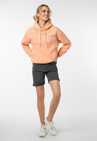 Perzikkleurige hoodie met trekkoorden en een voorzak, met witte grafische tekst. Draag het met opgerolde donkere shorts en witte sneakers.