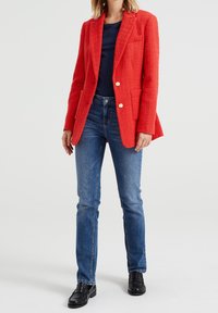 Blazer rosso con texture, rever a punta, due tasche frontali e bottoni bianchi, abbinato a jeans blu a gamba dritta e mocassini neri.