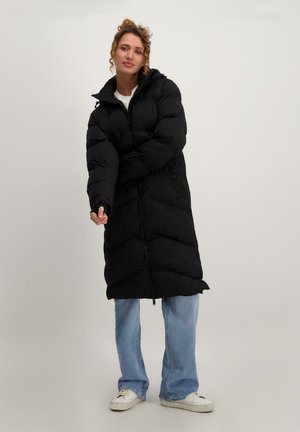 Zwarte pufferjas met een hoge kraag, lange lengte en gewatteerd design. Heeft een ritssluiting aan de voorkant en zakken, gedragen over lichtblauwe jeans en witte sneakers.