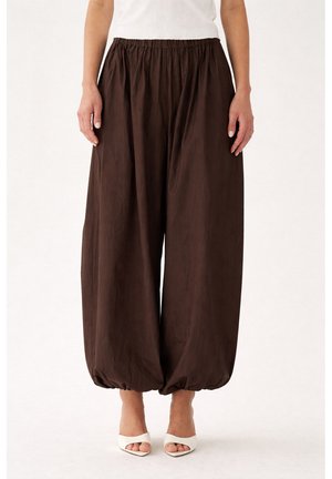 Løse brune bukser med samlet elastisk talje og manchetter, båret med hvide åbne hæle og en hvid ærmeløs top.