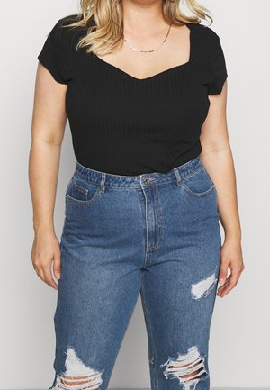 Kvinde iført en tætsiddende sort kortærmet top og højtaljede blå revnede jeans, stående mod en ensfarvet baggrund.
