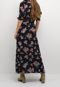 Robe maxi noire à imprimé floral avec des motifs roses et bleus, manches courtes retournées, ceinture à la taille et silhouette fluide. Portée avec des bottes à talons blocs beiges.