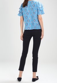 Niebieska bluzka kwiatowa z warstwowymi falbanami, noszona z czarnymi, dopasowanymi spodniami i granatowymi balerinami, podkreślająca lekką strukturę materiału.