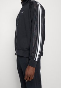 Chaqueta de entrenamiento negra de Nike con cierre a cremallera, cuello alto y acentos de malla texturizada con rayas blancas en las mangas.