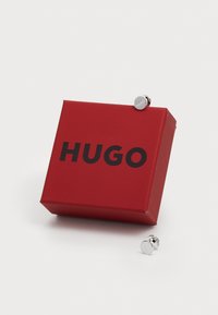 Röd presentask med "HUGO" i svart text, innehållande ett par silverfärgade runda manschettknappar med en polerad yta. Asken har en stilren design.
