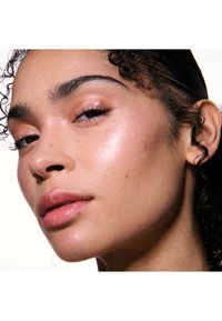 Close-up van een gezicht met een gladde, glanzende huid. Het onderwerp heeft donker krullend haar, zachte roze lippen en subtiele oogmake-up met gedefinieerde wenkbrauwen.
