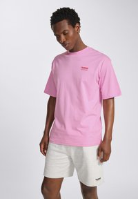 Lyserød bomulds T-shirt med korte ærmer, der har et lille rødt logo på venstre bryst, parret med lysegrå shorts. Blød tekstur og afslappet pasform.