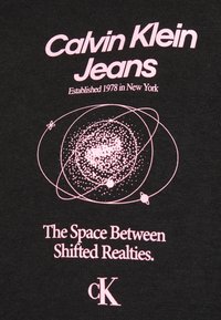 Svart t-shirt i tyg med rosa grafisk design. Texten inkluderar "Calvin Klein Jeans," etableringsår och en tagline med ett kosmiskt motiv.