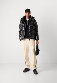 Giacca a vento nera lucida con cappuccio, maglione nero, pantaloni crema con strisce nere sui lati e sneakers nere. Porta una piccola borsa nera.