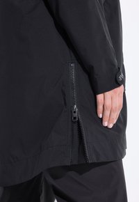 Giacca nera lunga con cerniera laterale parzialmente aperta, indossata da una persona che tiene la mano vicino alla cerniera, polsino della manica contrassegnato con testo.