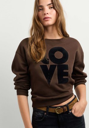 Giovane donna con lunghi capelli castano chiaro che indossa una felpa marrone con la scritta "LOVE" in grandi lettere nere e jeans scuri con cintura marrone.