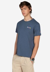 Camiseta de algodón azul con cuello redondo, mangas cortas y el texto "HARPER" en letras coloridas en el pecho. Textura suave y ajuste casual.